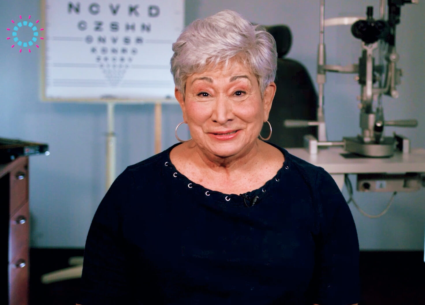 An elderly woman with short gray hair and a warm smile sits in an ophthalmology clinic. An eye chart and examination equipment are visible in the background.