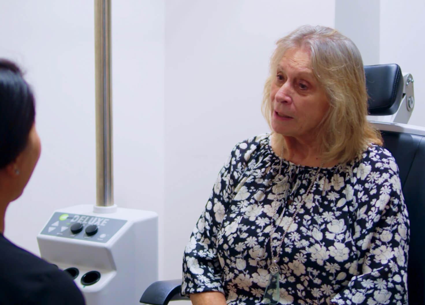 An older woman with blonde hair, wearing a floral blouse, sits in an ophthalmology exam chair, attentively speaking with a medical professional whose back is facing the camera.