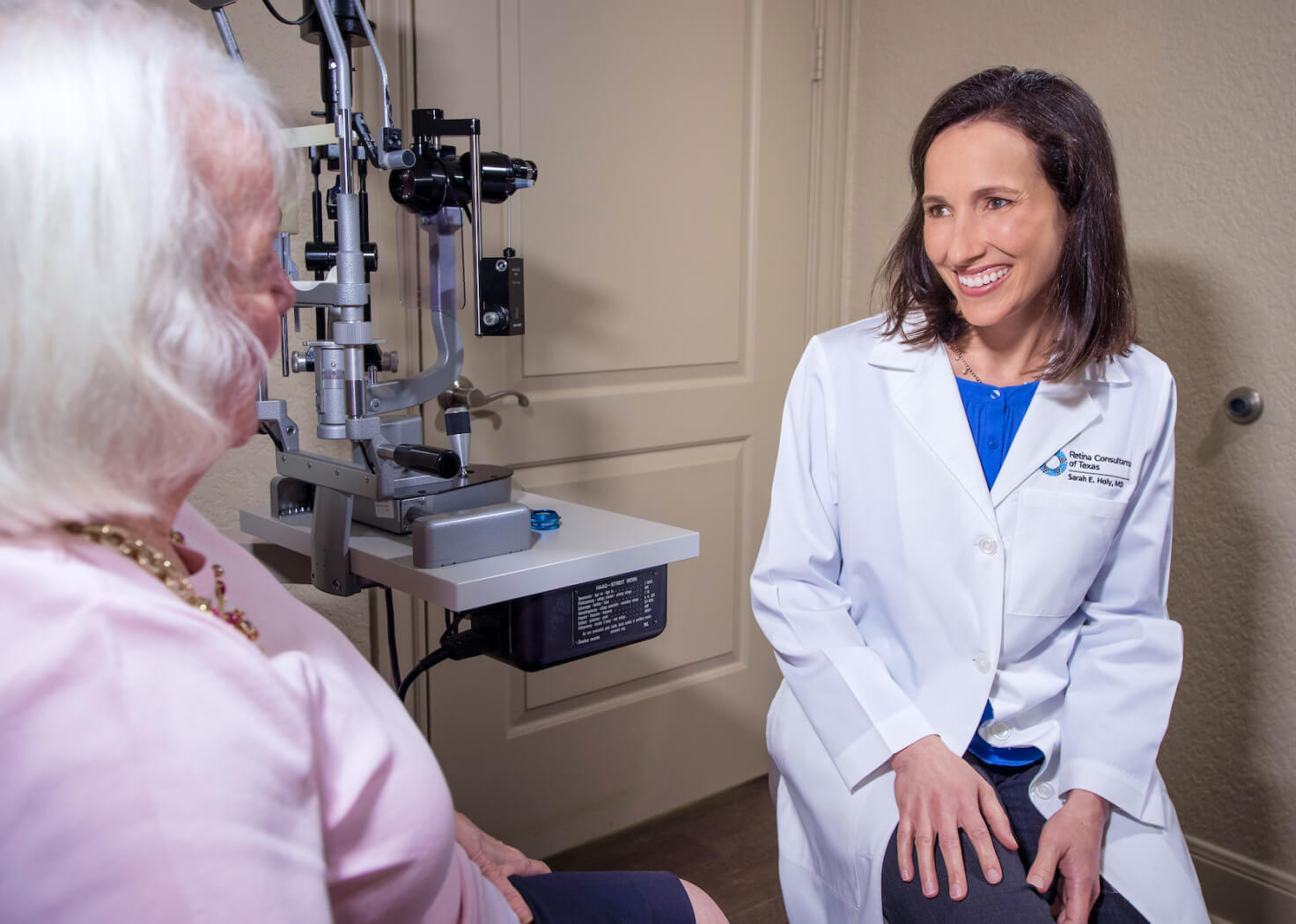 A smiling female doctor in a white coat sits and speaks with an elderly patient in an ophthalmology clinic