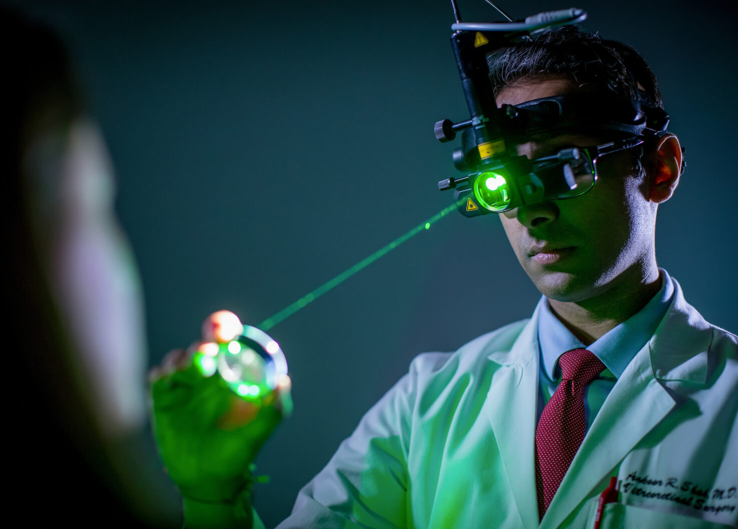 A doctor in a white coat and red tie performs an eye examination using a head-mounted ophthalmoscope and a green laser light in a dimly lit setting.