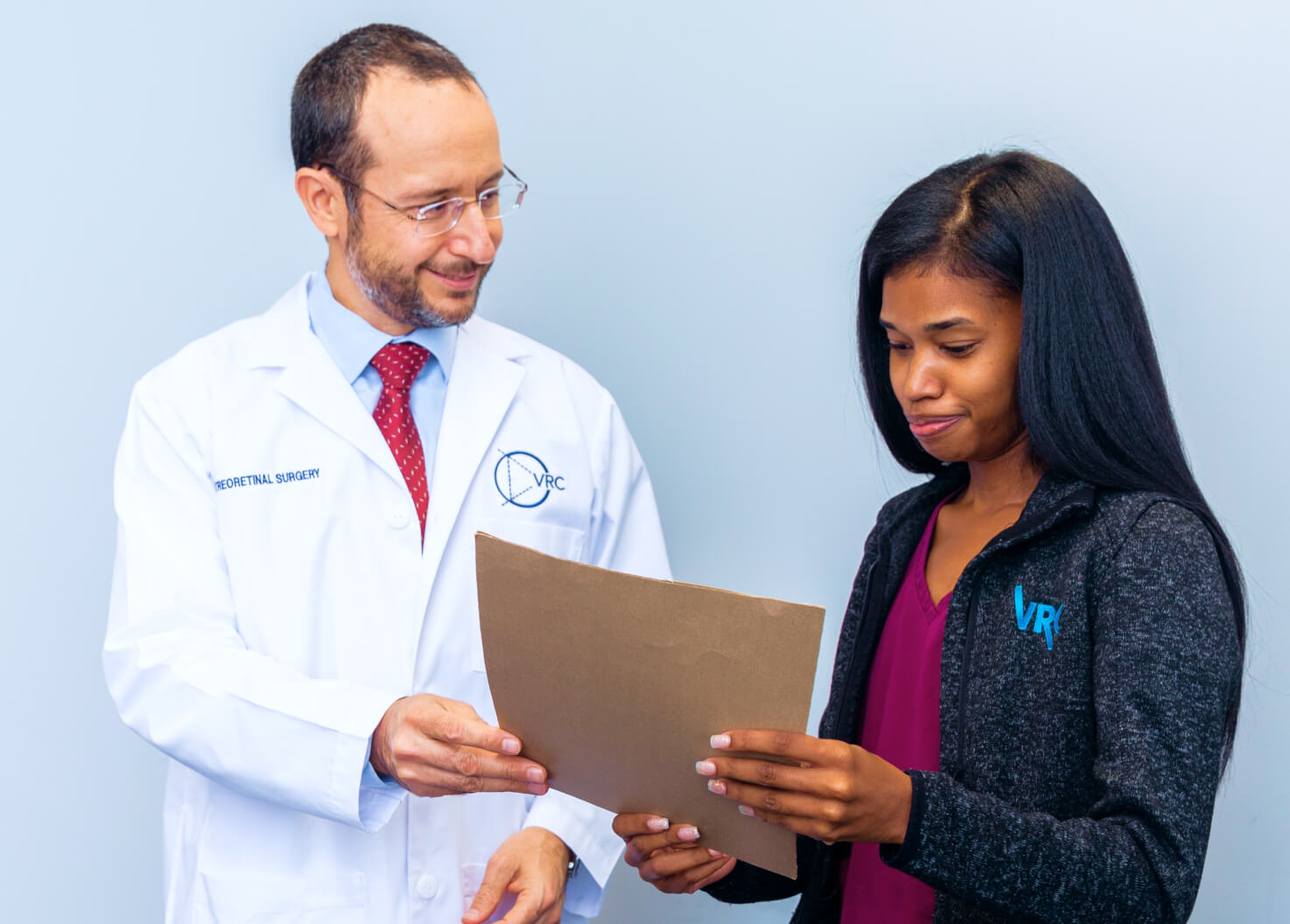 A doctor in a white coat and red tie discusses a file with a woman in a black jacket. They are both looking at the document against a plain light blue background.