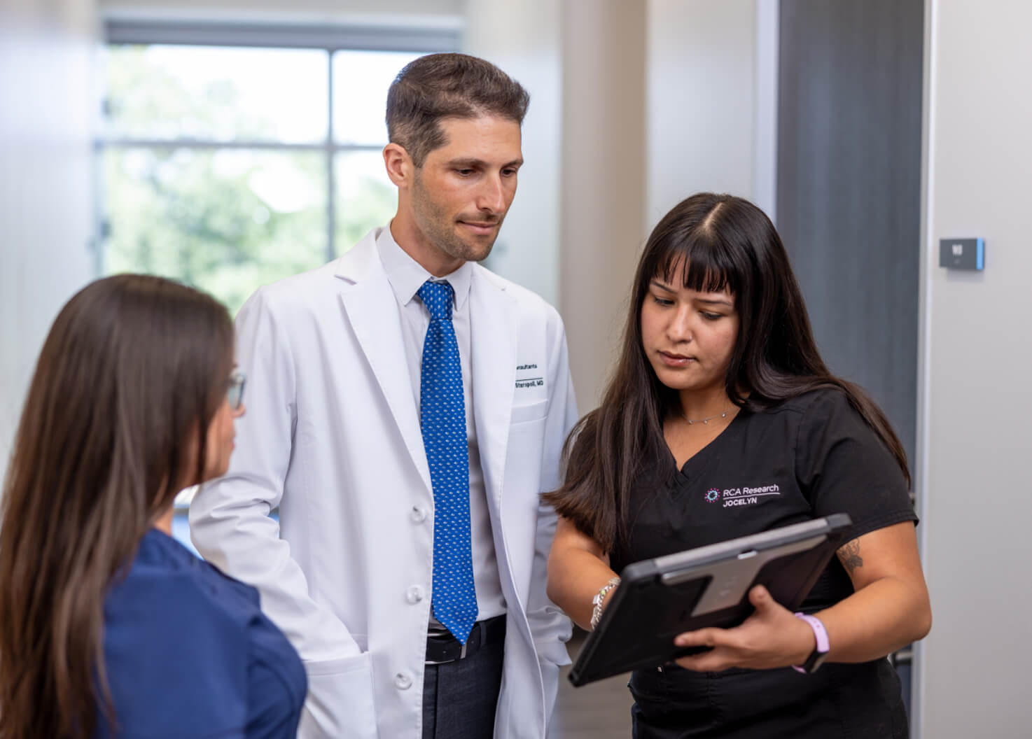 A doctor in a white coat and two medical professionals in scrubs discuss patient information while reviewing a tablet in a clinical hallway.