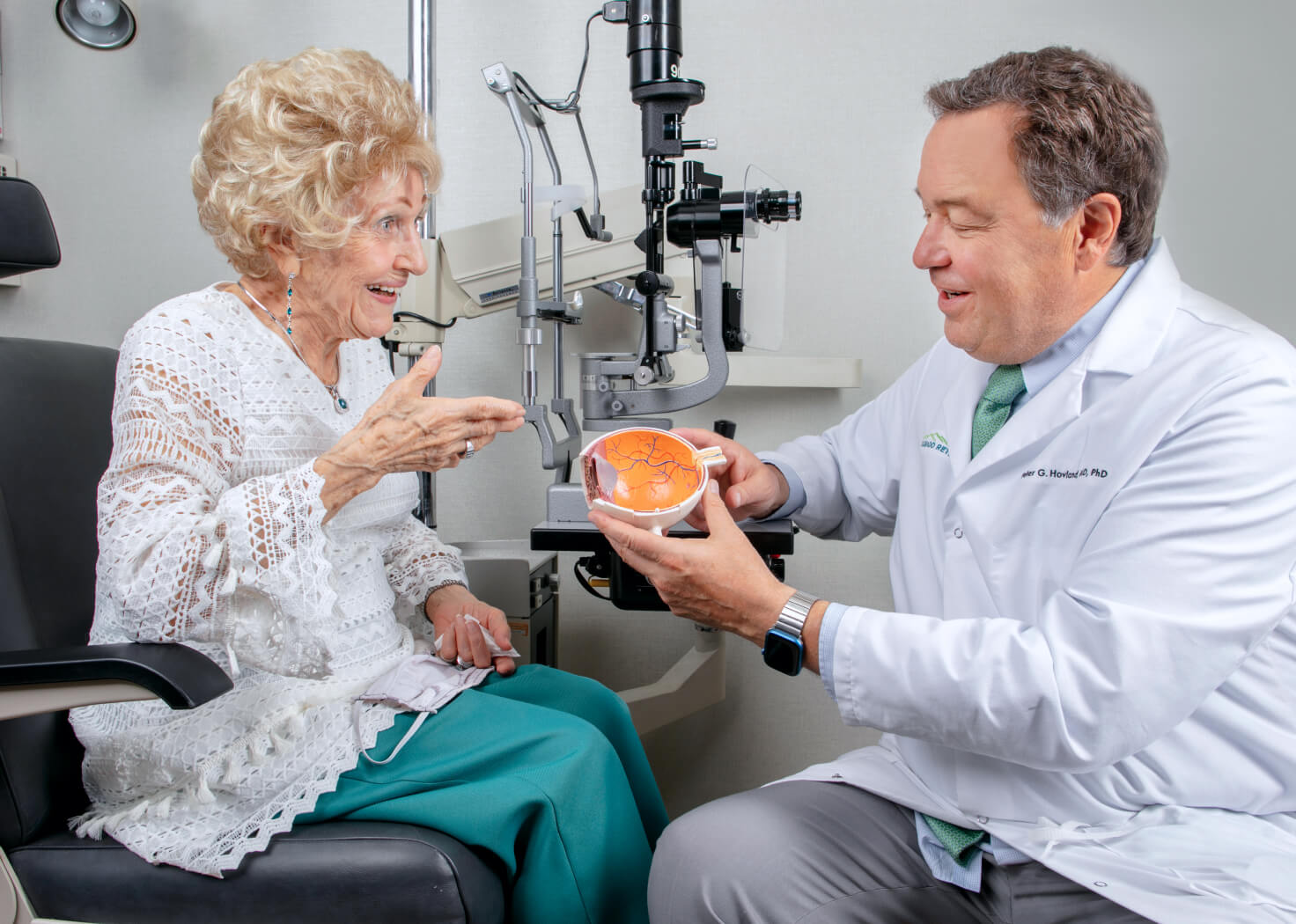A doctor in a white coat explains an eye condition to an elderly woman in an ophthalmology clinic, using a detailed anatomical model of the eye