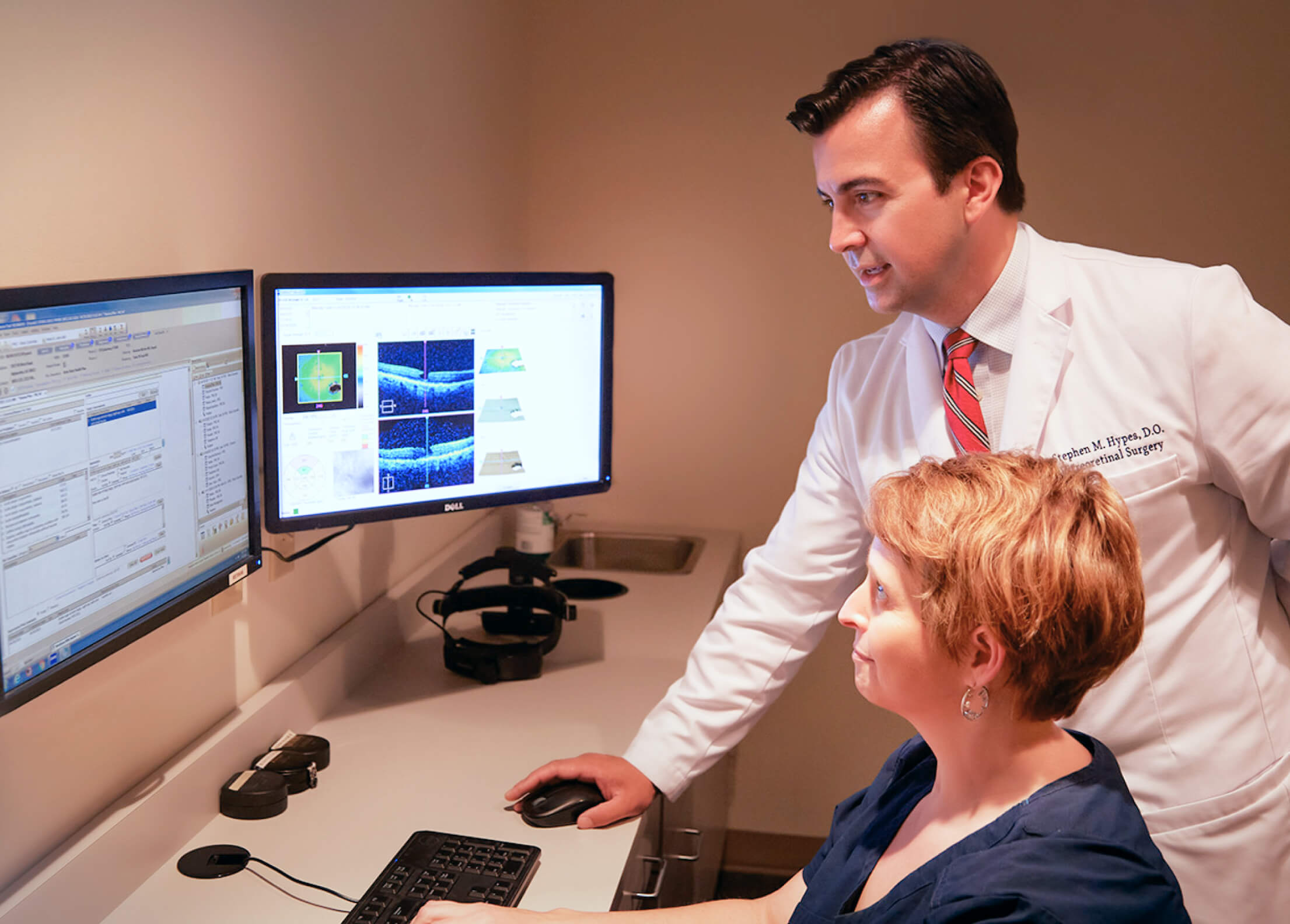 A doctor in a white coat with a name badge is assisting a colleague at a computer workstation.