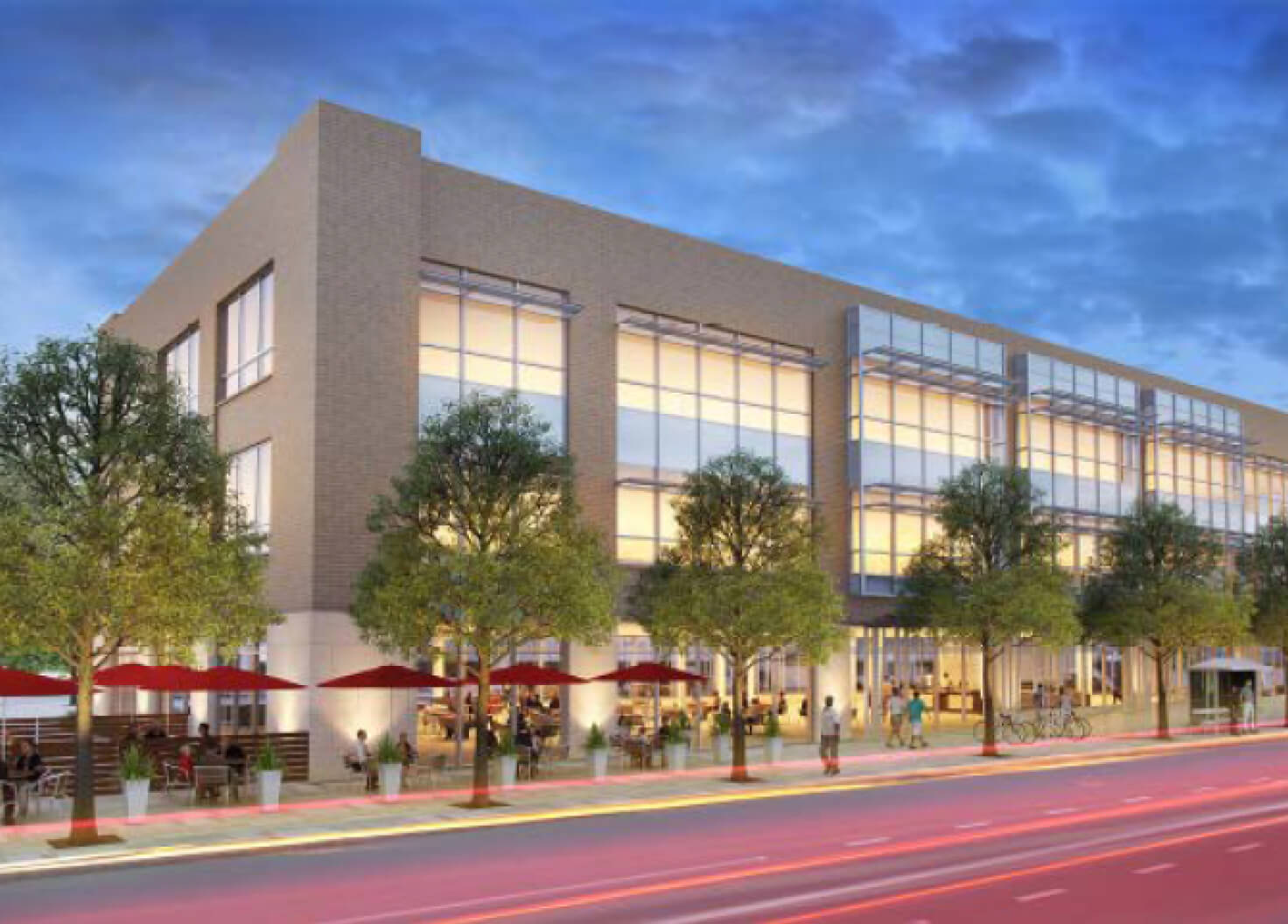 A modern three-story commercial building with large glass windows is illuminated at dusk. The street is lined with trees, and outdoor seating areas with red umbrellas are visible along the sidewalk.