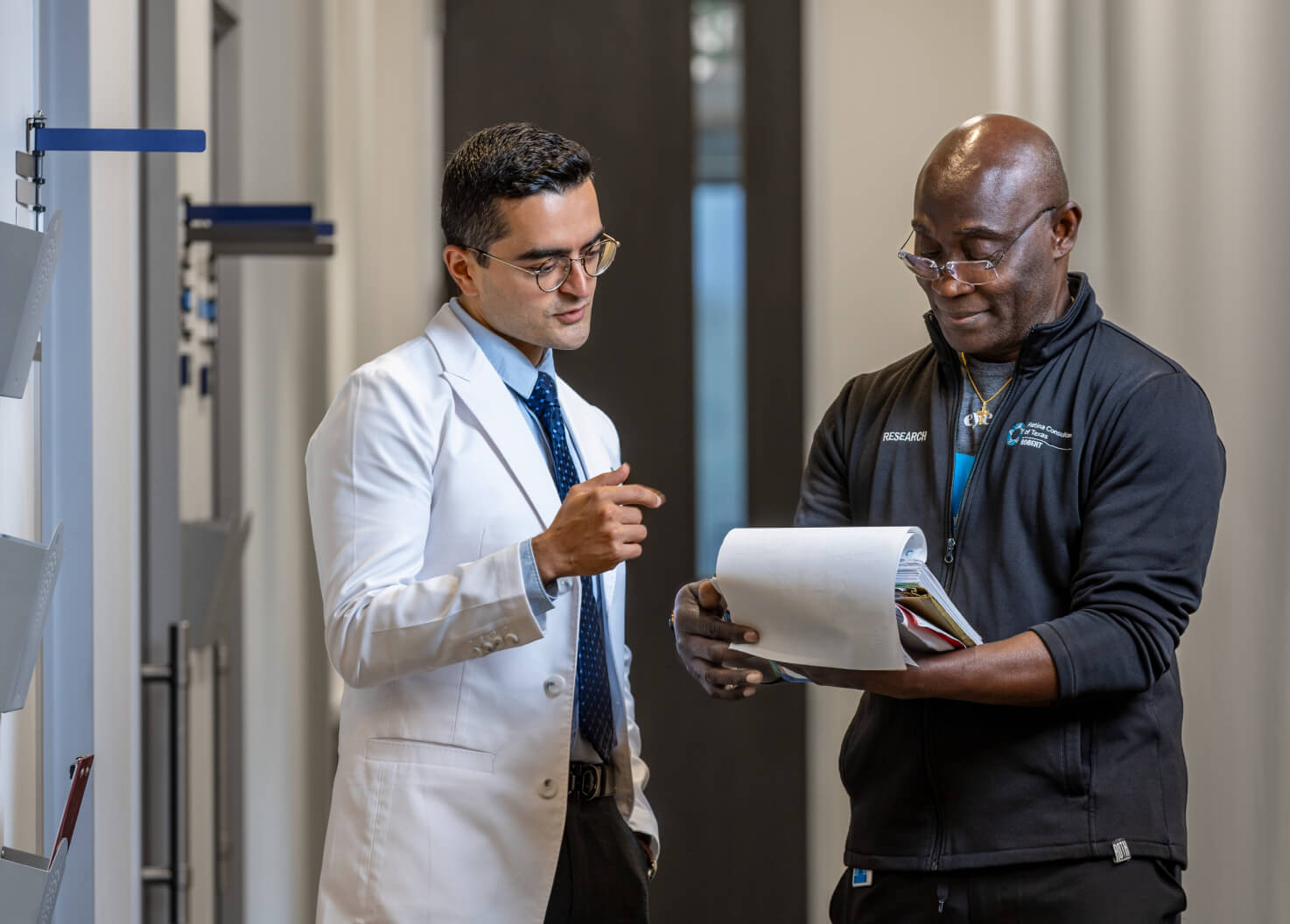 A doctor in a white coat discusses a document with a staff member in a black jacket. They are both engaged in conversation while reviewing paperwork in a professional setting.