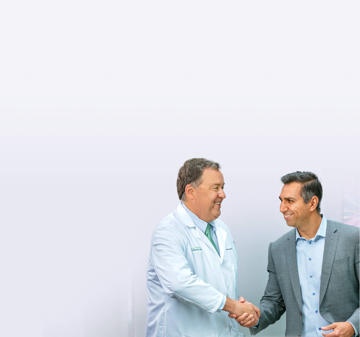 A doctor in a white coat and a man in a suit shake hands while smiling against a minimalistic, light gradient background.