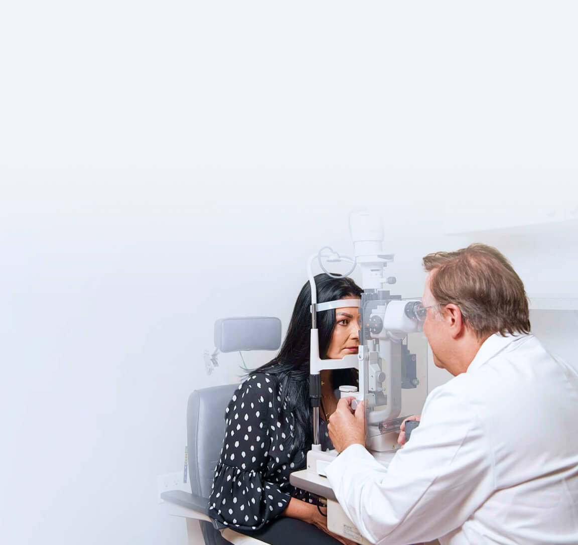 A doctor in a white coat performs an eye examination on a female patient using a slit lamp, against a minimalistic, light gradient background.